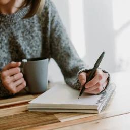 A woman holds a mug with her right hand and is about to write something in a notebook with her left